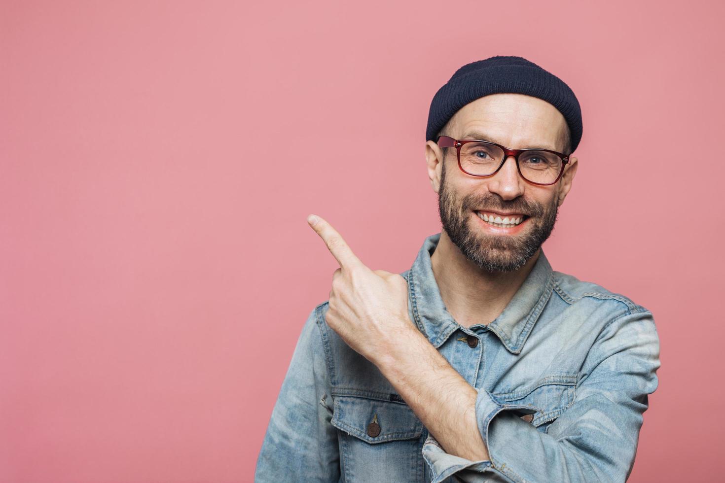 colpo orizzontale di allegro uomo barbuto con espressione felice, vestito con abiti alla moda, indossa occhiali, cappello e camicia di jeans, indicato con il dito anteriore nello spazio vuoto della copia per la tua pubblicità foto