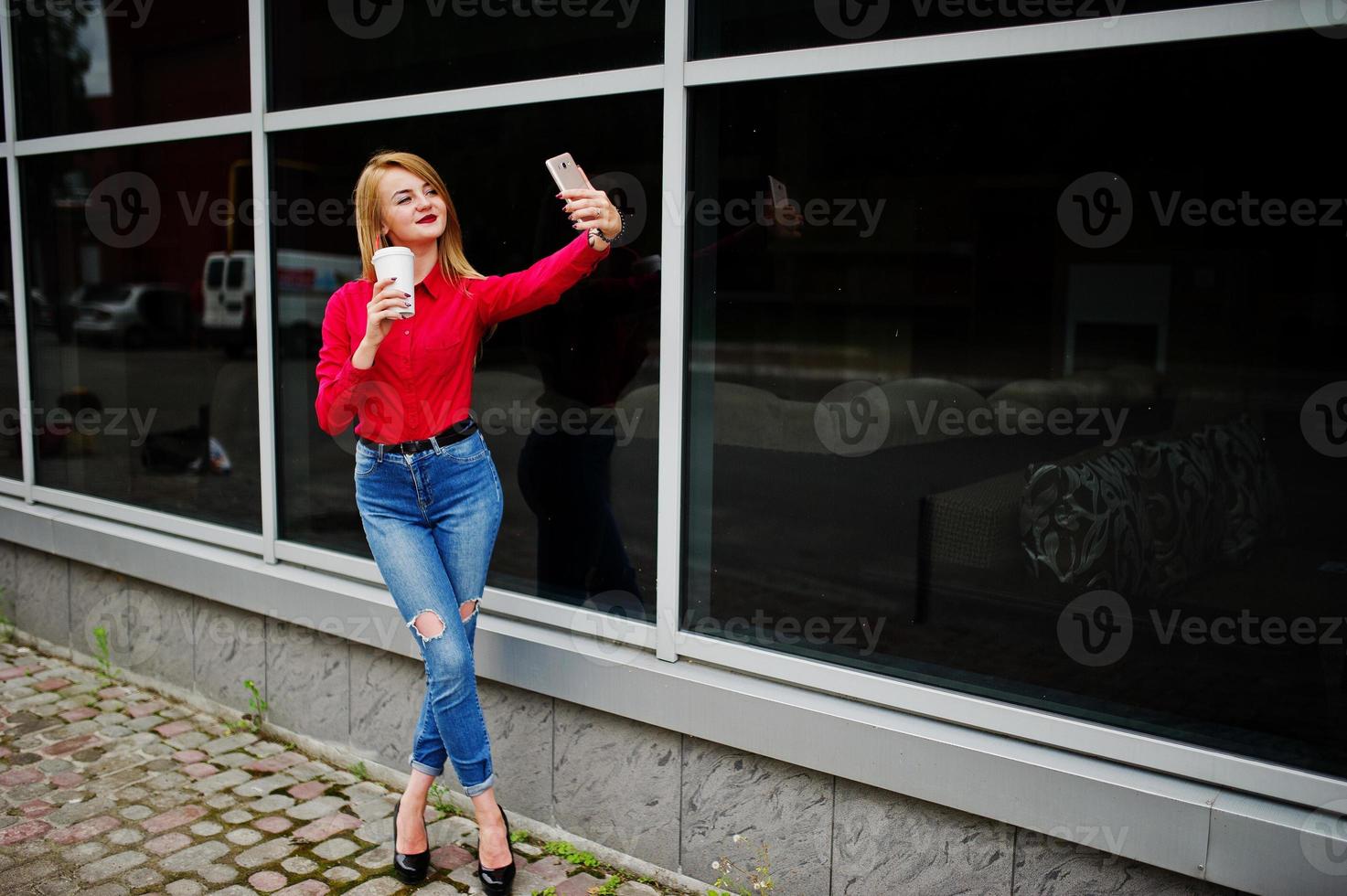 ritratto di una bella donna in camicetta rossa e jeans casual che si fa selfie sul cellulare e tiene una tazza di caffè fuori dall'enorme centro commerciale. foto