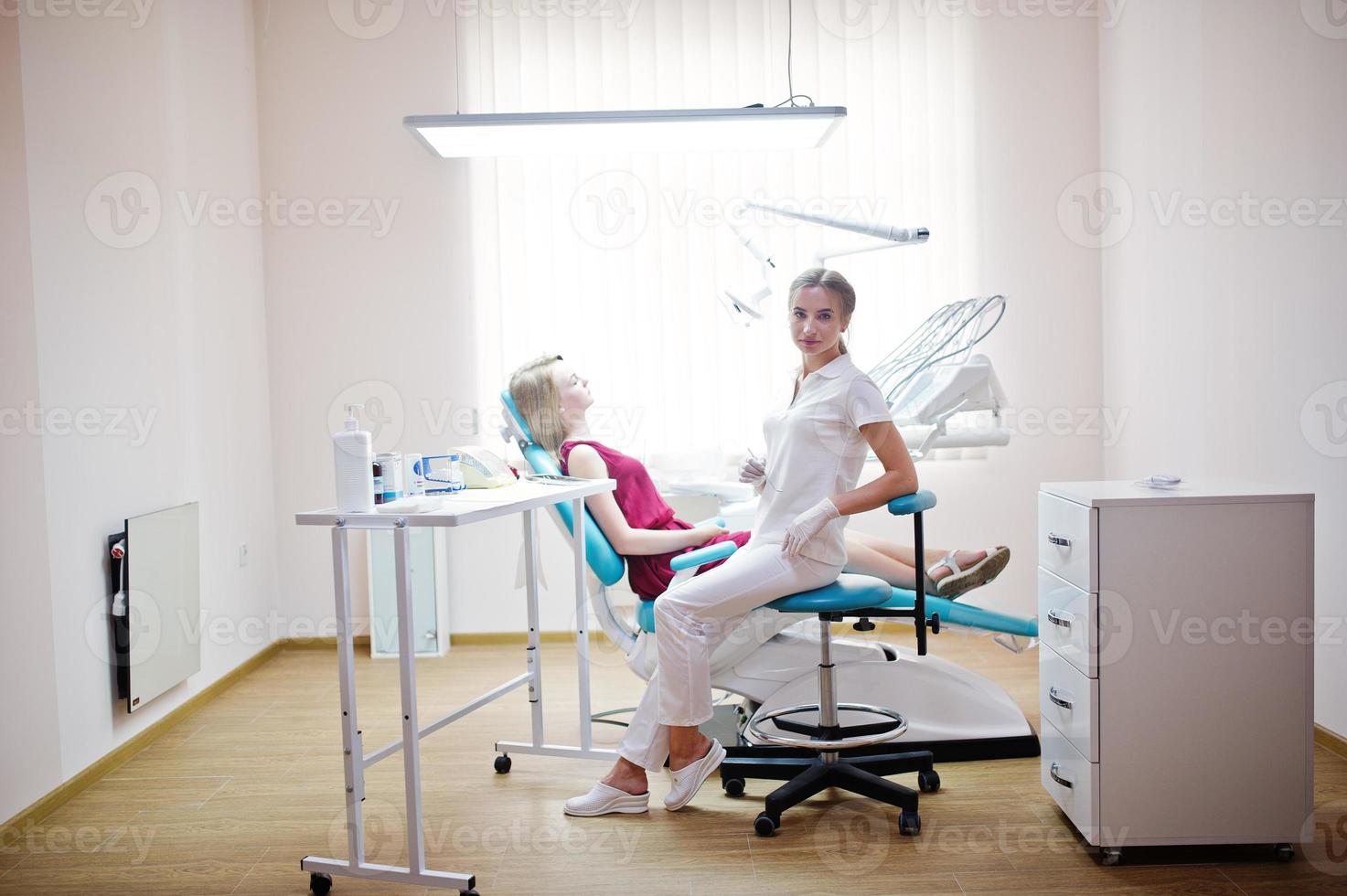 bella dentista femminile in posa e sorridente con il suo adorabile paziente sdraiato su una poltrona odontoiatrica in abito rosso-viola. foto