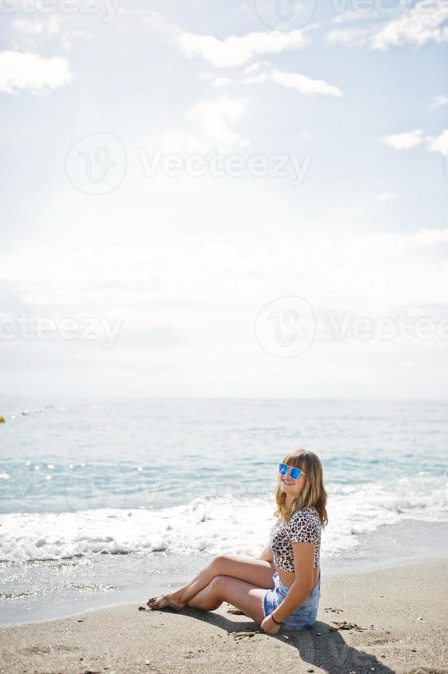 bellissima modella che si rilassa su una spiaggia di mare, indossa jeans corti, camicia leopardata e occhiali da sole. foto