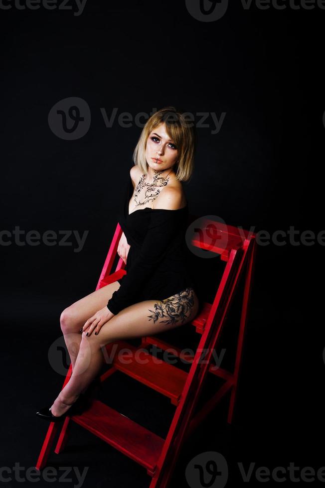 ritratto in studio di ragazza bionda con trucco originale sul collo e tatuaggio sulla coscia, indossa un abito nero su sfondo scuro, seduto su una scala rossa. foto