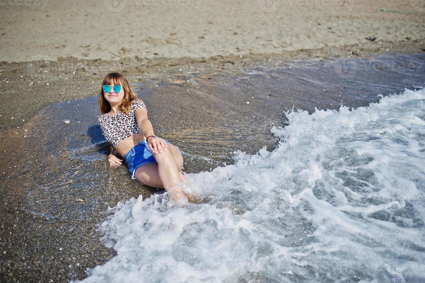 bellissima modella che si rilassa su una spiaggia di mare, indossa jeans corti, camicia leopardata e occhiali da sole. foto