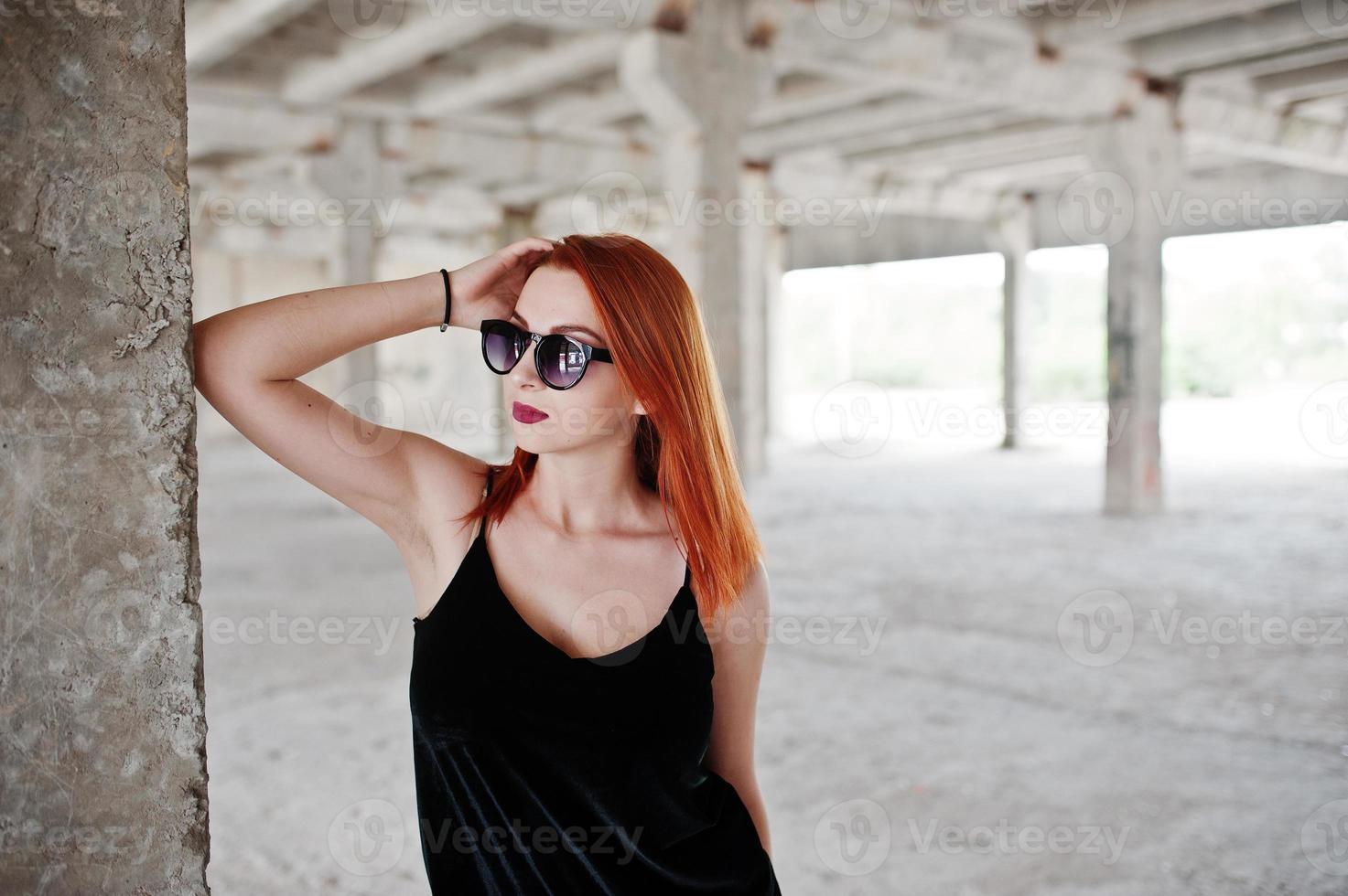 la ragazza alla moda dai capelli rossi in occhiali da sole indossa in nero, contro il luogo abbandonato. foto