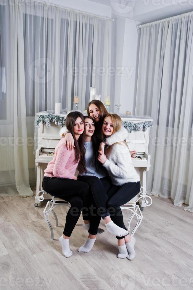 quattro ragazze carine amiche indossano maglioni caldi e pantaloni neri contro il vecchio pianoforte con decorazioni natalizie nella stanza bianca. foto