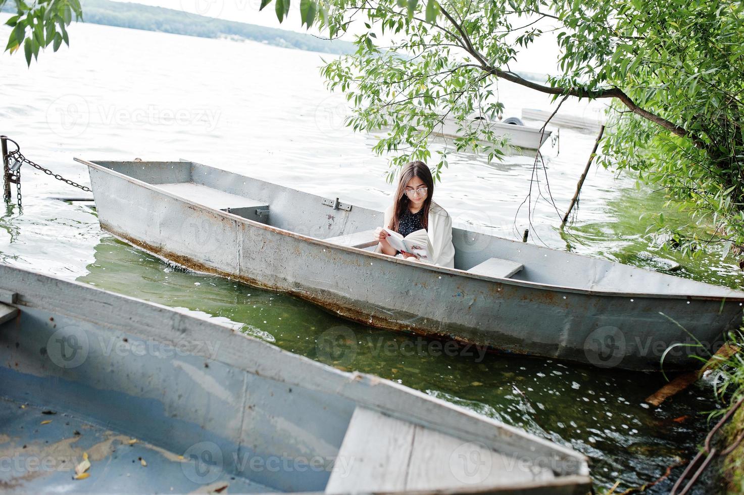 ritratto di una donna attraente che indossa un abito nero a pois, uno scialle bianco e occhiali che legge un libro in una barca su un lago. foto