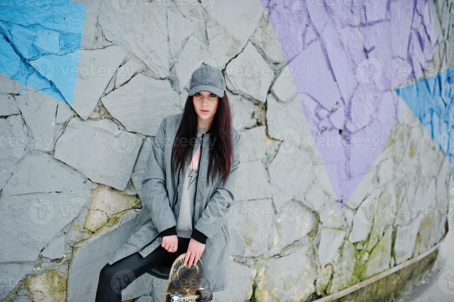 elegante ragazza bruna con berretto grigio, casual street style con skate board in una giornata invernale contro la parete colorata. foto