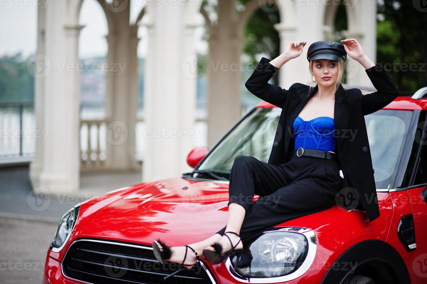 ritratto di bella bionda modella donna moda sexy in berretto e in tutto nero con trucco luminoso sedersi sul cofano della city car rossa. foto