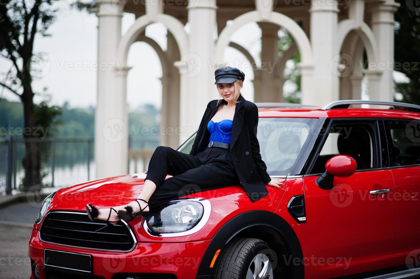 ritratto di bella bionda modella donna moda sexy in berretto e in tutto nero con trucco luminoso sedersi sul cofano della city car rossa. foto