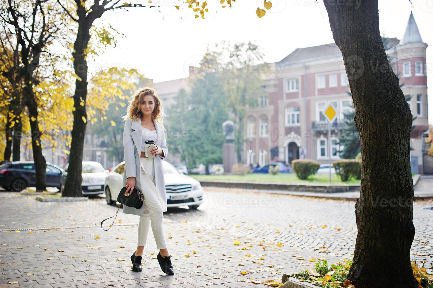 elegante modello biondo riccio ragazza indossare su bianco con una tazza di caffè a portata di mano in posa in autunno strada della città contro l'auto bianca. foto