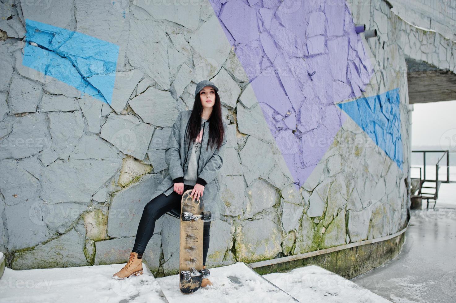 elegante ragazza bruna con berretto grigio, casual street style con skate board in una giornata invernale contro la parete colorata. foto