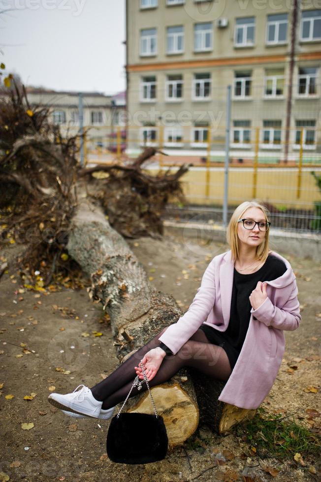 ragazza bionda con occhiali e cappotto rosa, tunica nera seduta su un albero tagliato. foto