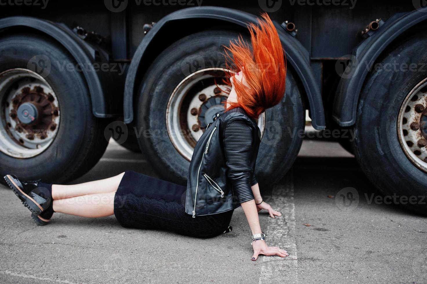 ragazza elegante dai capelli rossi indossa in nero, seduta contro grandi ruote di camion. foto