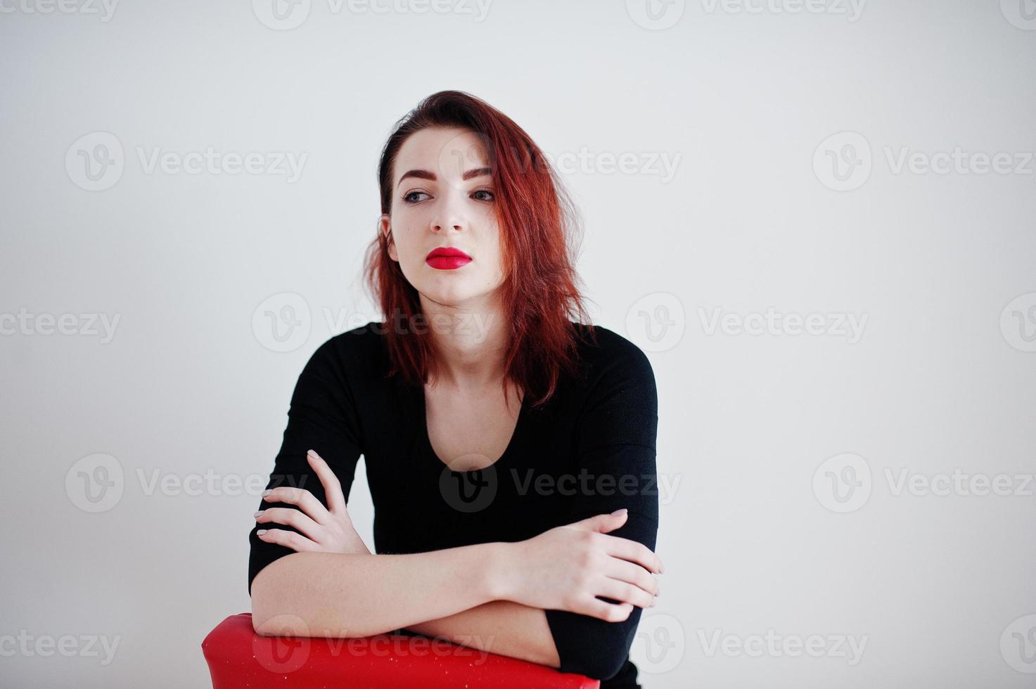 ragazza dai capelli rossi su tunica nera seduta su una sedia rossa contro il muro bianco in una stanza vuota. foto