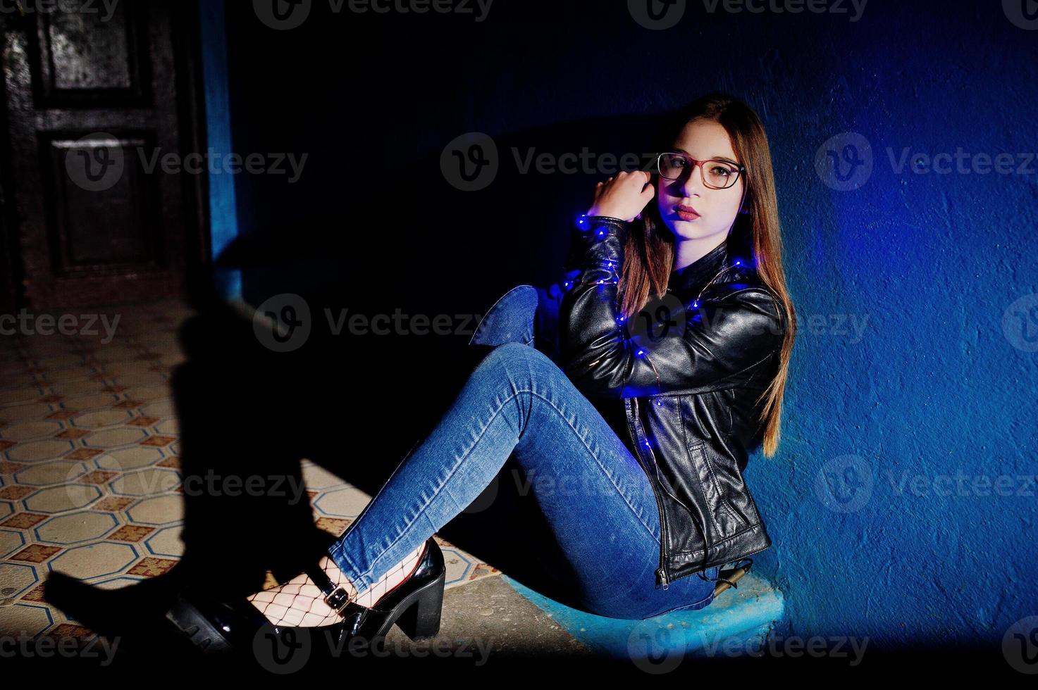 ritratto notturno di ragazza modello indossa occhiali, jeans e giacca di pelle, con ghirlanda blu su di lei. foto