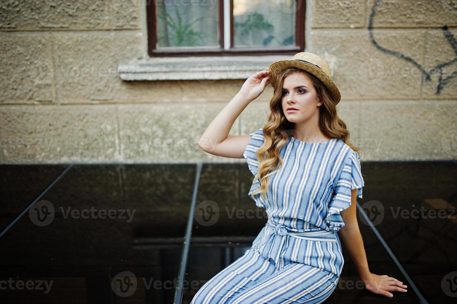 ritratto di una favolosa giovane donna che indossa tuta a righe e cappello seduto su una superficie nera lucida accanto all'edificio. foto