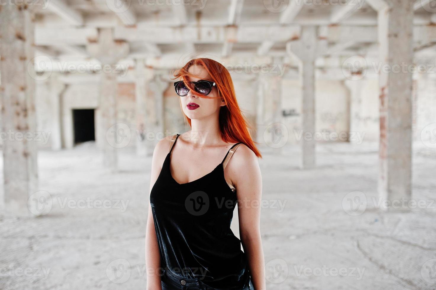 la ragazza alla moda dai capelli rossi in occhiali da sole indossa in nero, contro il luogo abbandonato. foto