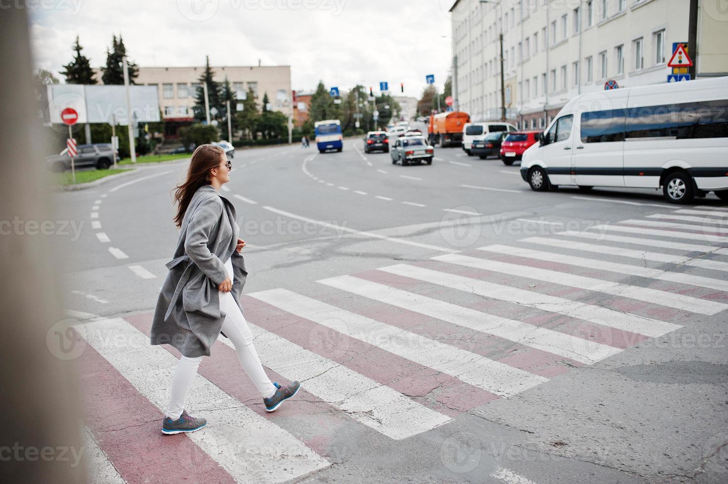 ragazza in cappotto grigio con occhiali da sole e borsetta che cammina sul passaggio pedonale. foto