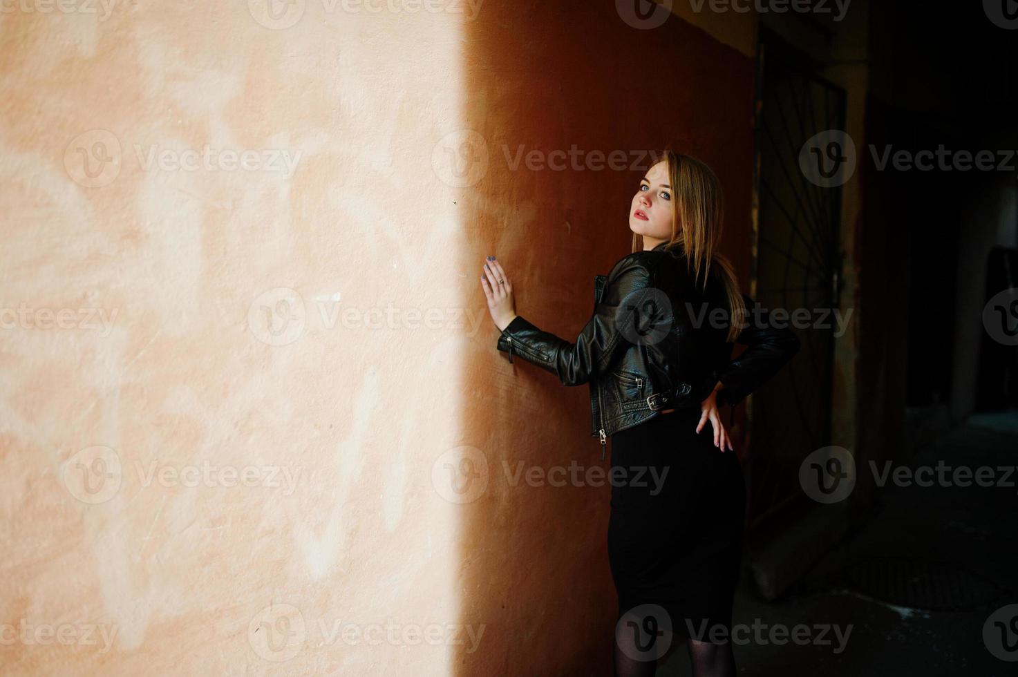 elegante ragazza bionda indossare su giacca di pelle nera in posa per le strade del vecchio muro di sfondo della città con le ombre. foto