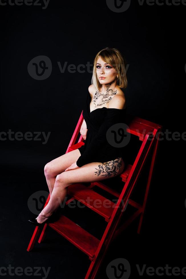 ritratto in studio di ragazza bionda con trucco originale sul collo e tatuaggio sulla coscia, indossa un abito nero su sfondo scuro, seduto su una scala rossa. foto