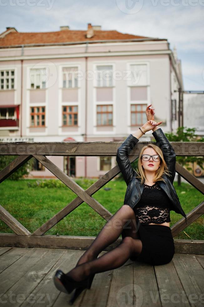 elegante ragazza bionda indossa una giacca di pelle nera in posa per le strade della città seduta su legno. foto