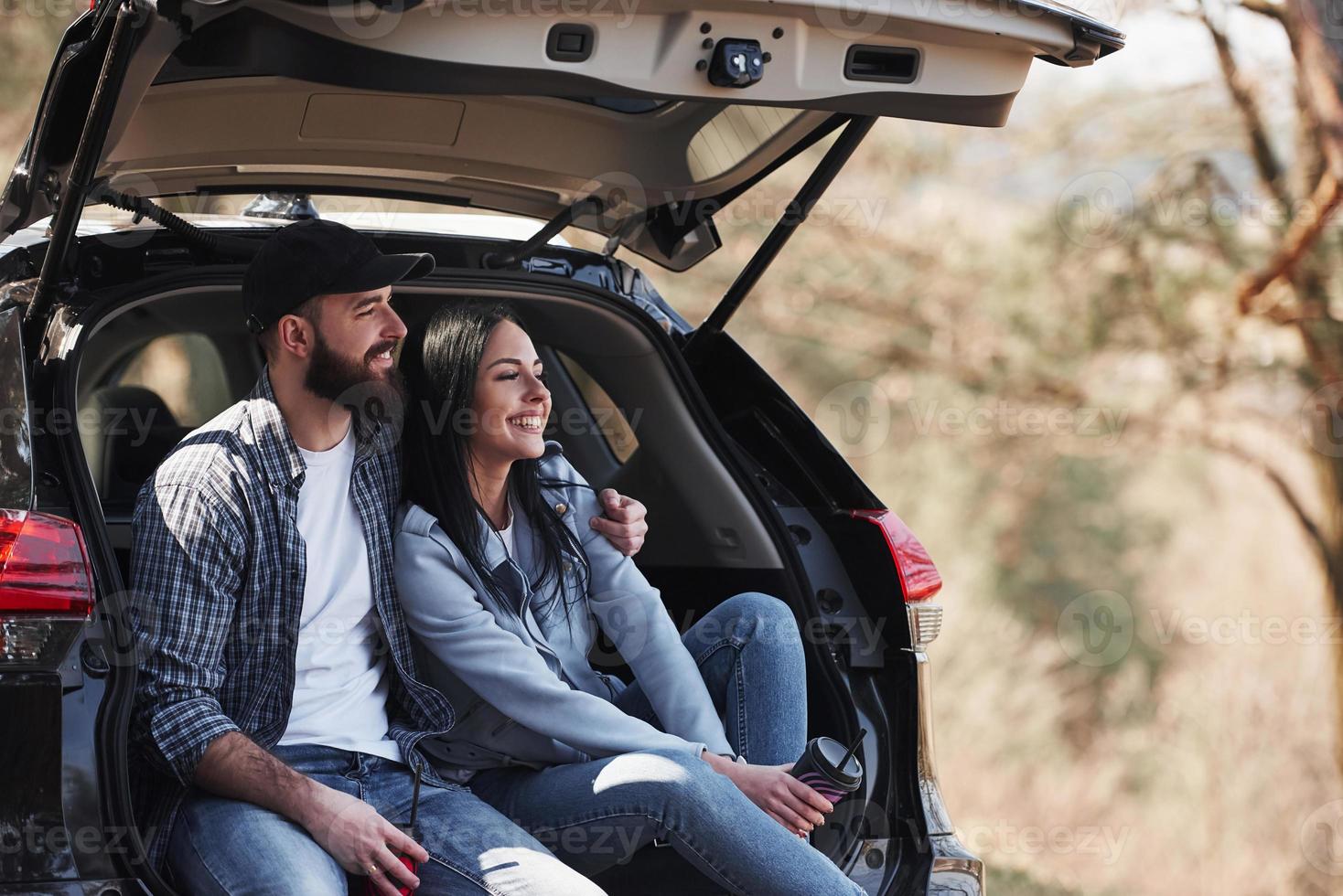 sorrisi a trentadue denti. seduto sulla parte posteriore dell'automobile. godersi la natura. la coppia è arrivata nella foresta con la loro auto nera nuova di zecca foto