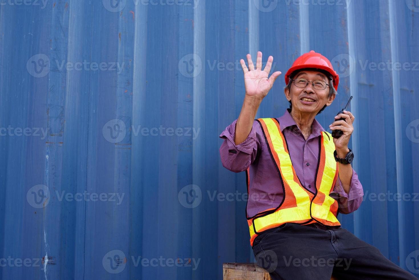 un anziano anziano ingegnere lavoratore asiatico che indossa giubbotto di sicurezza e casco in piedi e in possesso di walkie talkie radiofoniche presso il cantiere logistico di container di carico. foto