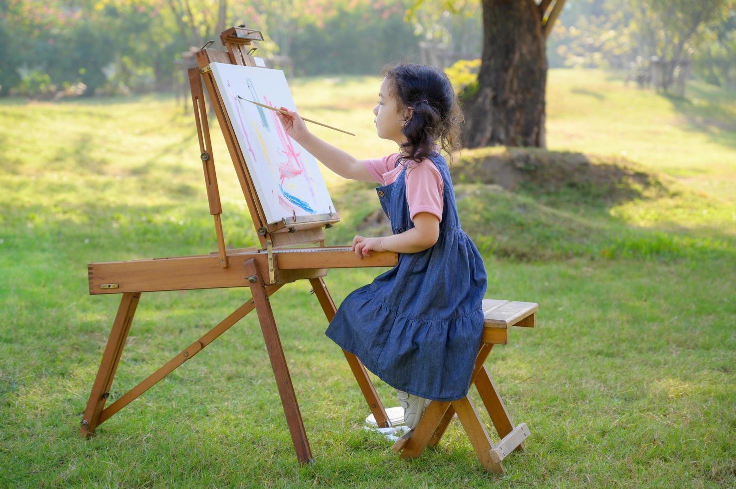 una bambina è seduta sulla panca di legno e dipinta sulla tela posta su un leggio da disegno foto