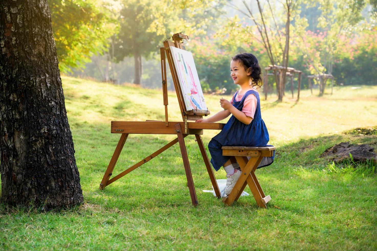 una bambina è seduta sulla panca di legno e dipinta sulla tela posta su un leggio da disegno foto