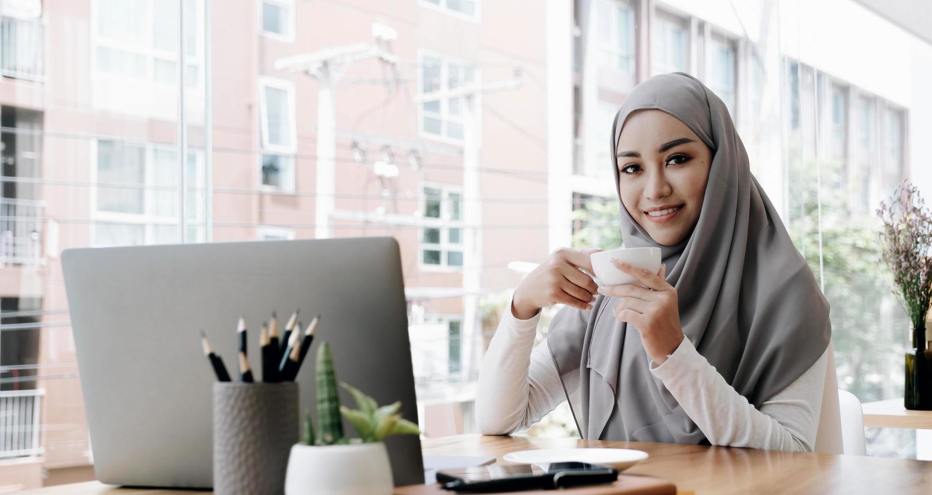 attraente e allegra lavoratrice musulmana asiatica o studentessa universitaria con hijab che lavora a distanza presso la caffetteria, tenendo una tazza di caffè. foto