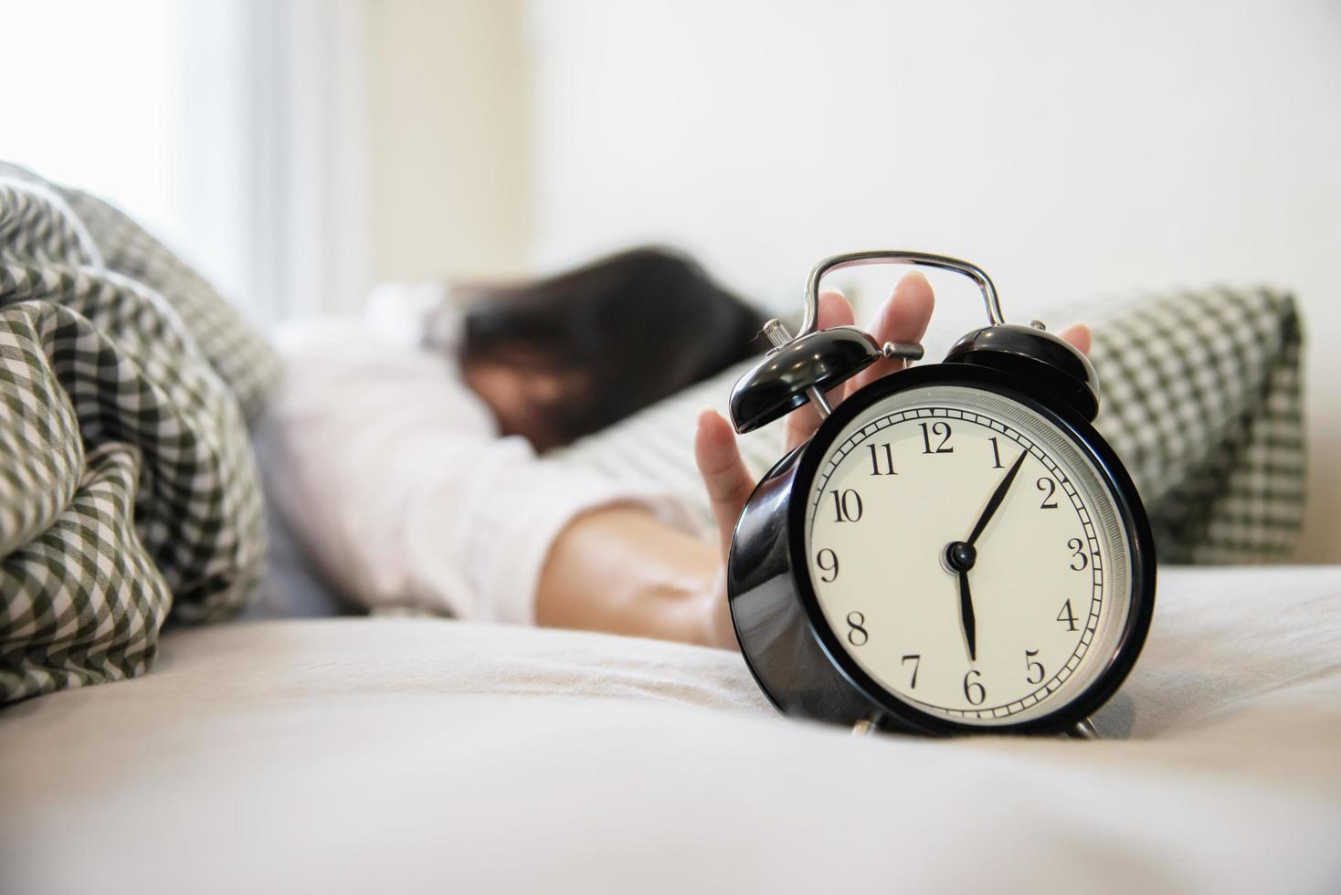 donna assonnata che raggiunge tenendo la sveglia al mattino con un risveglio tardivo - concetto di vita quotidiana a casa foto