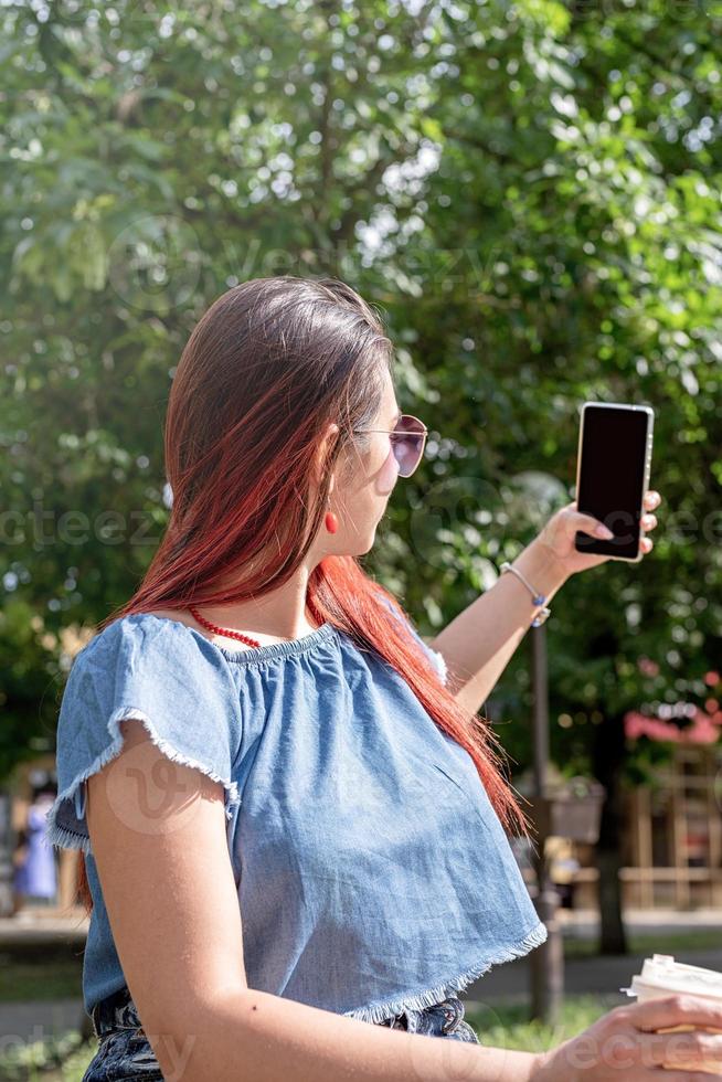 donna alla moda allegra con i capelli rossi che beve caffè al parco, prendendo selfie, schermo mockup foto