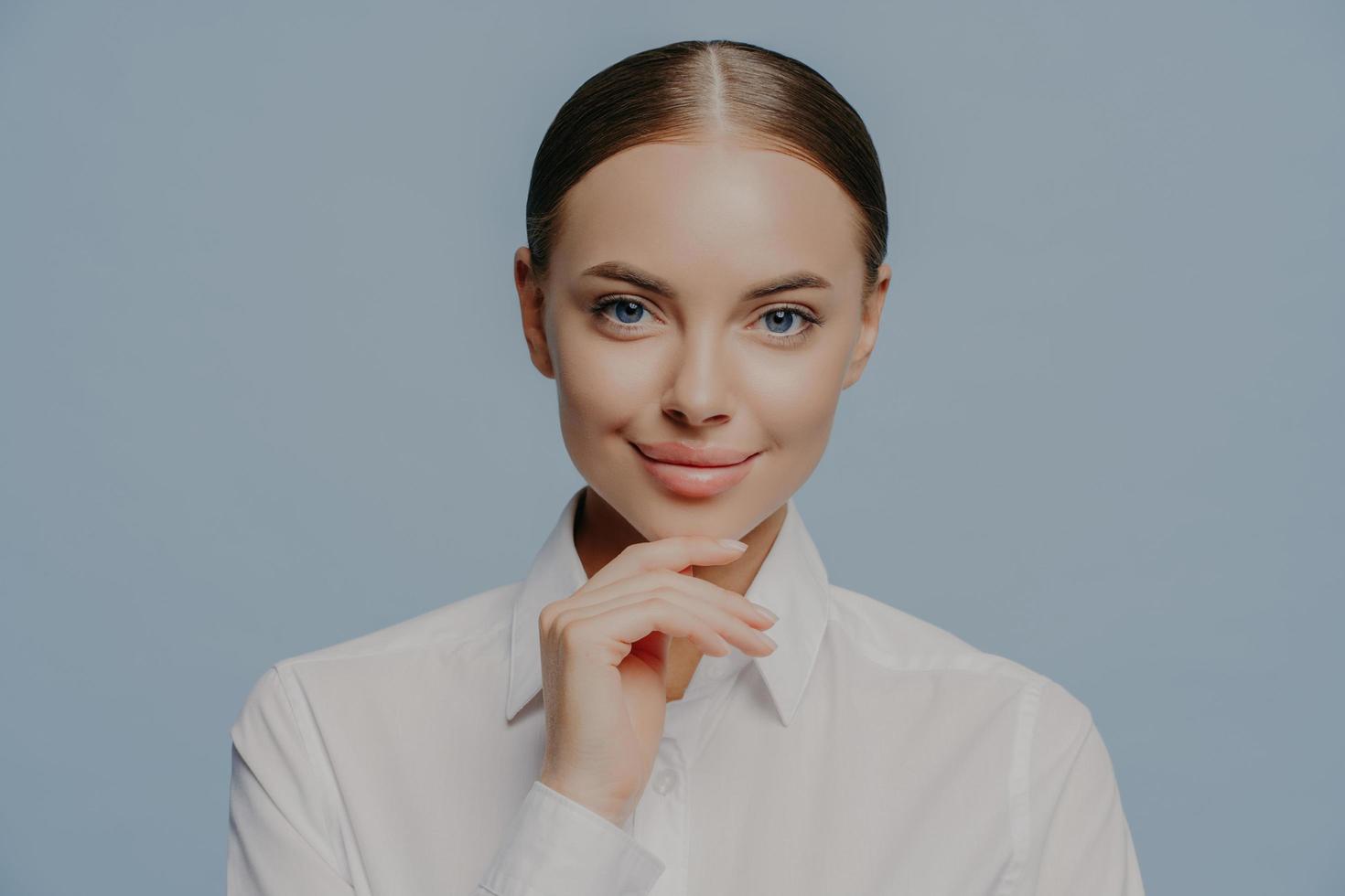 La donna d'affari attraente e sicura di sé tiene il mento e guarda direttamente la fotocamera, indossa un'elegante camicia bianca, ha una pelle sana e un trucco naturale, isolata su sfondo blu. donne, affari, carriera foto