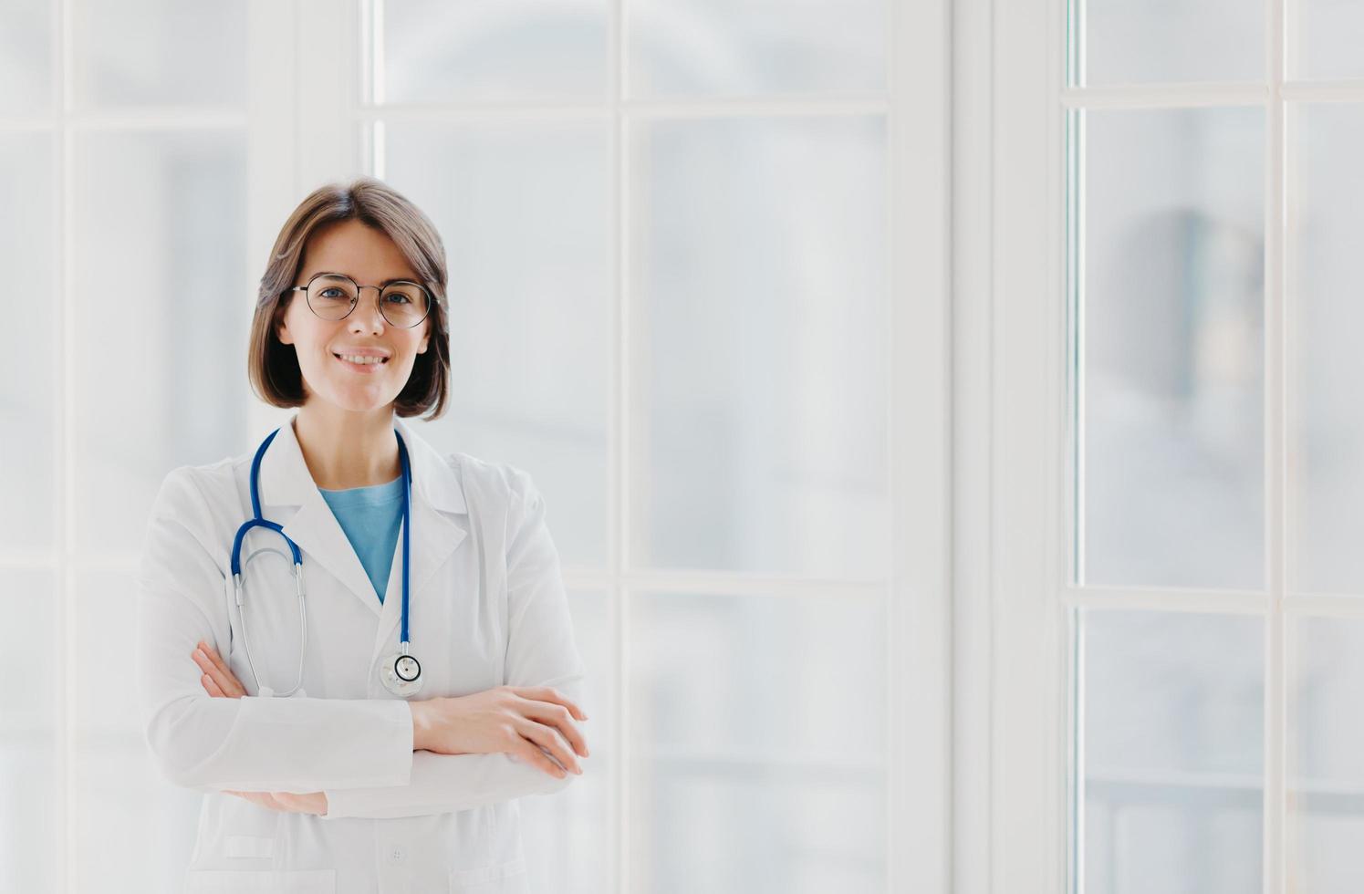 il medico professionista sicuro di sé sta con le braccia incrociate, indossa un camice medico bianco con uno stetoscopio, pensa al lavoro in modo positivo, posa contro una grande finestra. concetto di assistenza sanitaria e occupazione foto