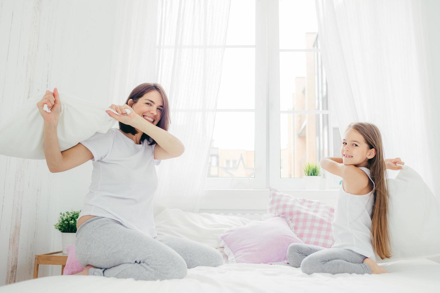 il ritratto di un bambino felice tiene il cuscino, litiga insieme a sua madre, si diverte in camera da letto, si gode il tempo libero, ha buone relazioni. la madre attraente in pigiama alleva sua figlia foto