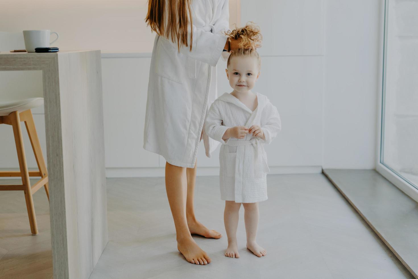 adorabile ragazza riccia carina posa vicino alla madre in morbido accappatoio bianco. la mamma irriconoscibile pettina i capelli della figlia fa la coda di cavallo a piedi nudi in un appartamento moderno. concetto di cura dei capelli e bambini foto