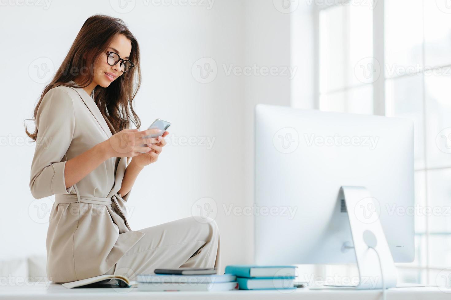 inquadratura orizzontale di bella giovane donna in abiti eleganti, ha un sorriso gentile, controlla il feed delle notizie tramite uno smartphone moderno, si riposa dopo un duro lavoro, posa all'interno dell'ufficio sul desktop, pareti bianche foto