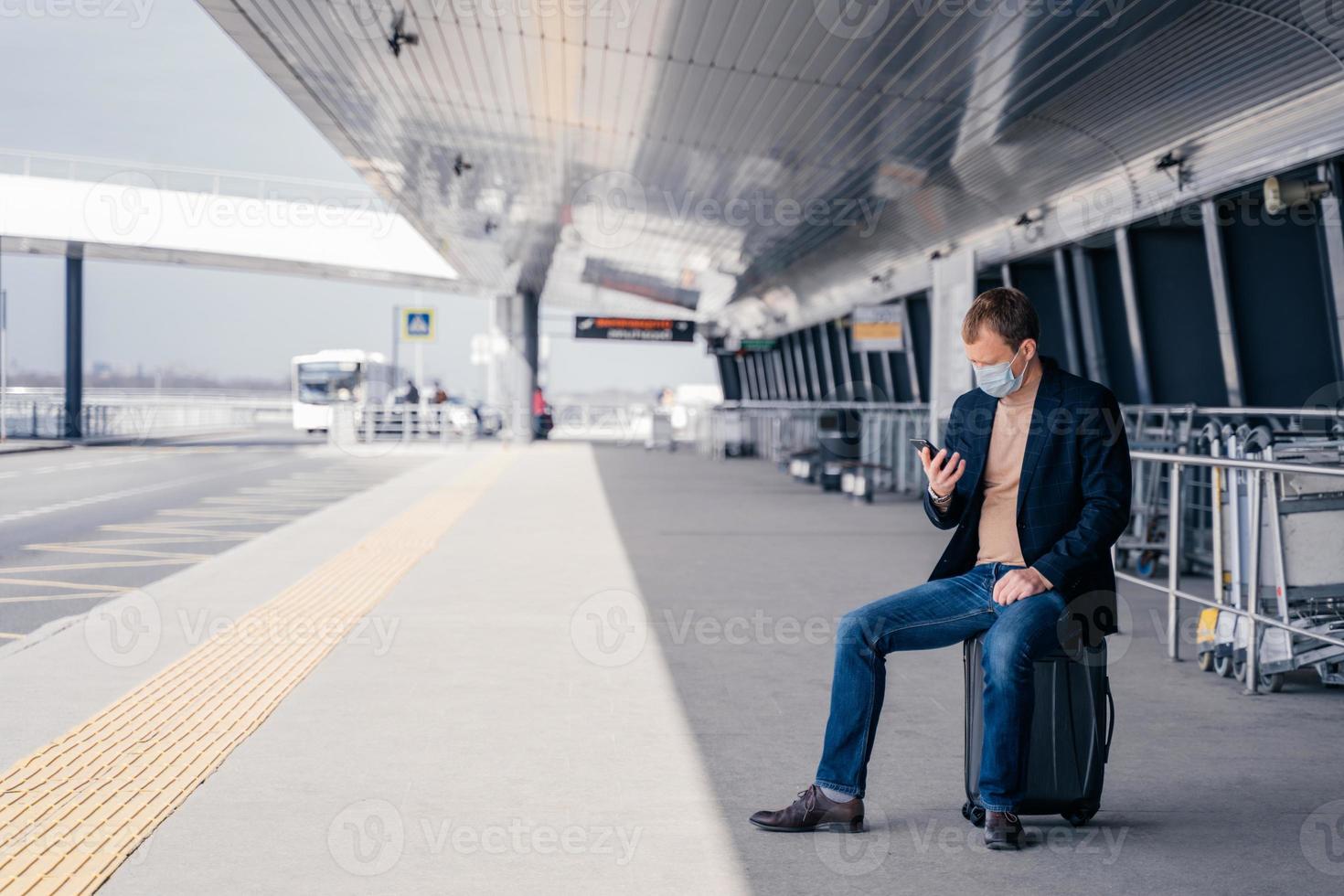 l'uomo europeo si siede alla valigia sulla stazione del binario degli autobus, cerca di trovare l'orario dei trasporti online su smartphone, attende il trasporto, viaggia durante una pericolosa situazione pandemica, indossa una maschera medica foto