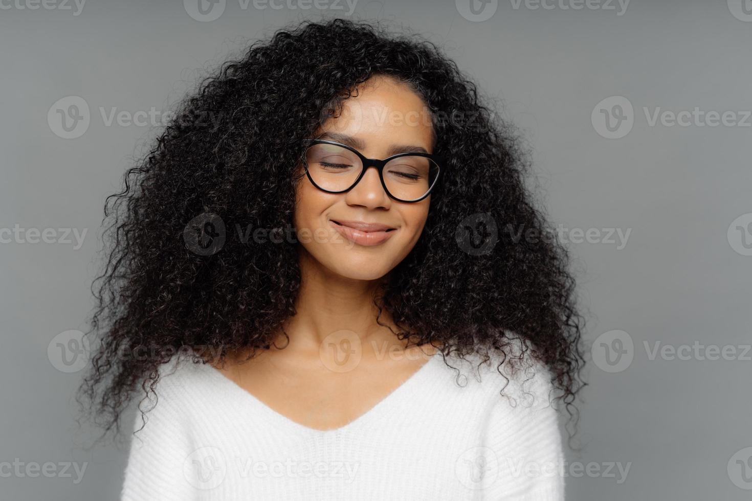 immagine ravvicinata di una donna sorridente compiaciuta con capelli folti e crespi, tiene gli occhi chiusi, sorride dolcemente, indossa occhiali ottici, immagina qualcosa di piacevole, vestita con un maglione bianco, isolata su un muro grigio scuro foto