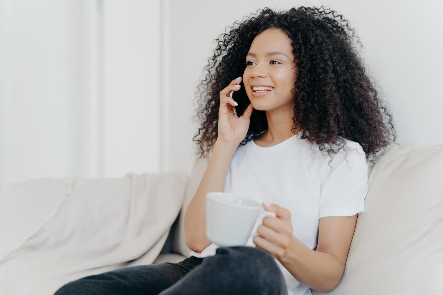 inquadratura laterale di una donna bruna etnica positiva ha una conversazione telefonica, beve una bevanda calda, tiene una tazza bianca, si siede su un comodo divano in soggiorno, sente piacevoli notizie, sorride felicemente foto