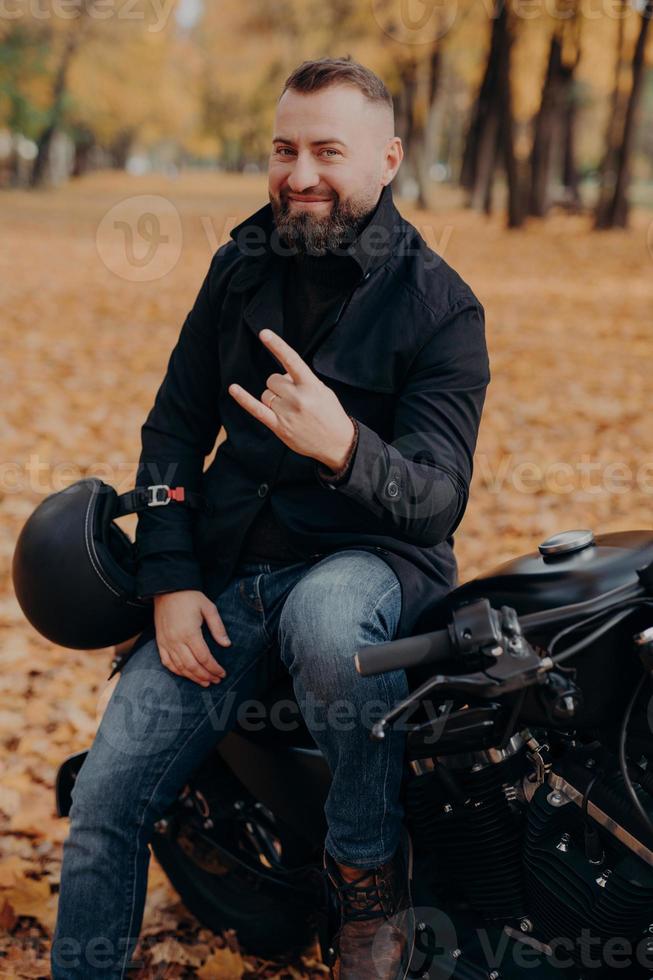 bel motociclista maschio con la barba tiene il casco, fa il gesto del clacson con le dita, si sente fresco, indossa un cappotto nero e jeans, si siede su una moto veloce sullo sfondo del parco autunnale. corridore spensierato foto