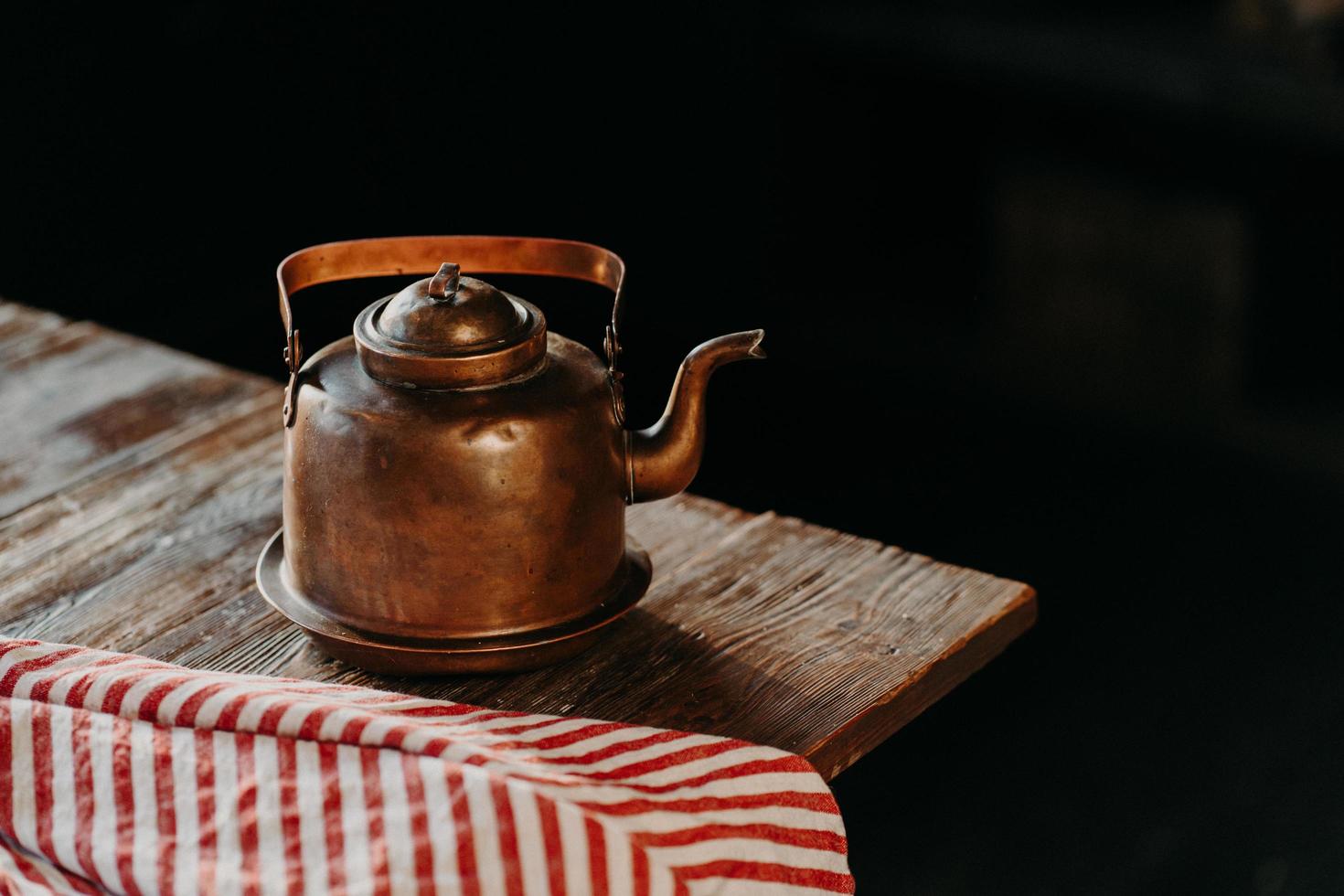 colpo orizzontale di bollitore antico su tavola di legno vintage. vecchie stoviglie. teiera di rame metallico su sfondo scuro asciugamano rosso vicino. messa a fuoco selettiva foto