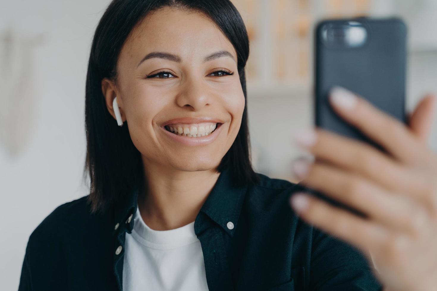 sorridente amichevole giovane donna che indossa l'auricolare comunica durante la videochiamata tenendo in mano lo smartphone foto