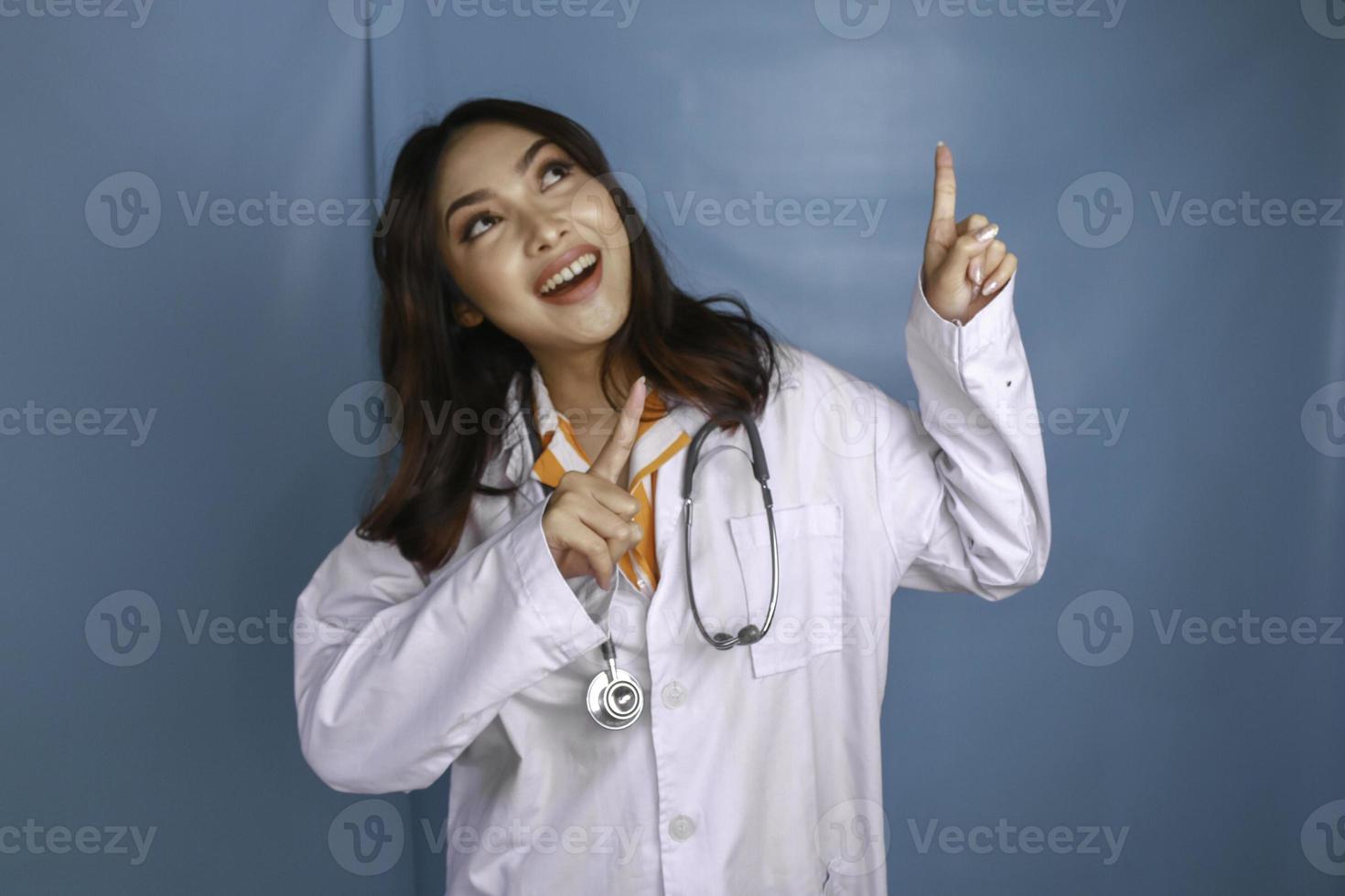 ritratto di una giovane donna asiatica medico, un medico sorride e punta verso l'alto in uno spazio di copia isolato su sfondo blu foto