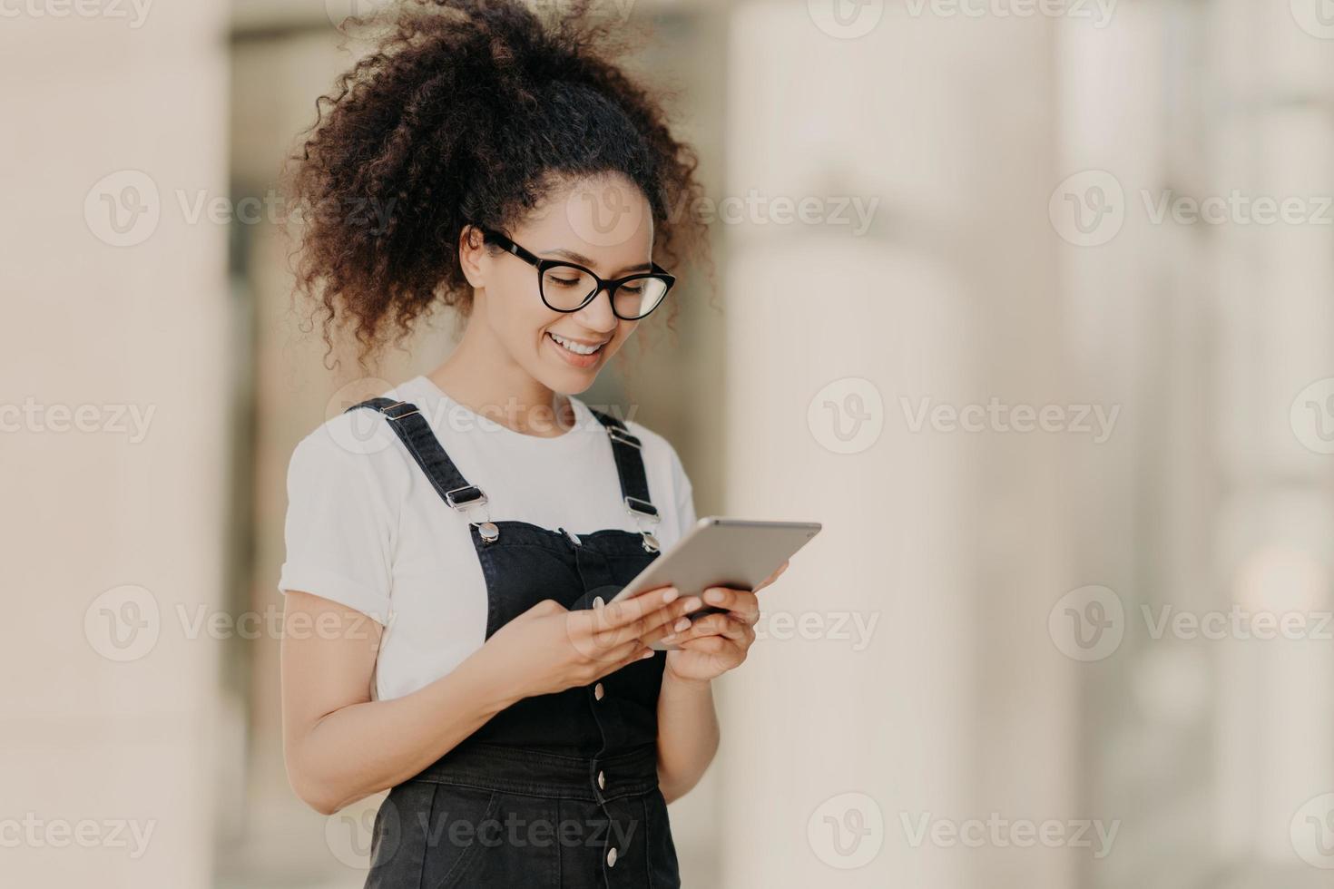 bella ragazza dai capelli ricci guarda lo schermo del tablet digitale con un sorriso, legge del testo o un libro elettronico, indossa occhiali per la correzione della vista, maglietta bianca e salopette, gode di internet ad alta velocità foto