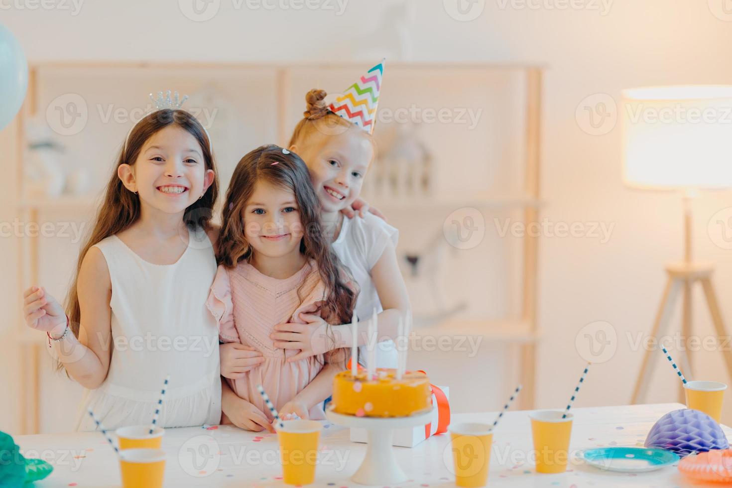 un'immagine orizzontale di tre amici felici si abbracciano e hanno espressioni divertenti, posano vicino a un tavolo festivo con una torta al coperto. tre ragazze festeggiano il compleanno insieme, si divertono, vengono a festeggiare foto