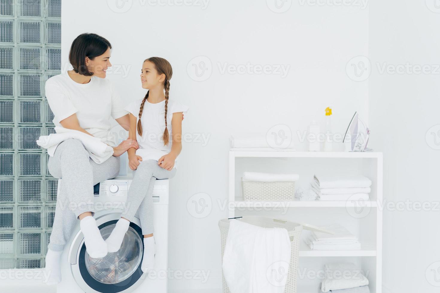 la madre premurosa parla con la figlia, posa sulla lavatrice, circondata da lenzuola bianche, lava insieme, stando in lavanderia a casa. la casalinga adulta felice ringrazia la ragazza adorabile per l'aiuto foto