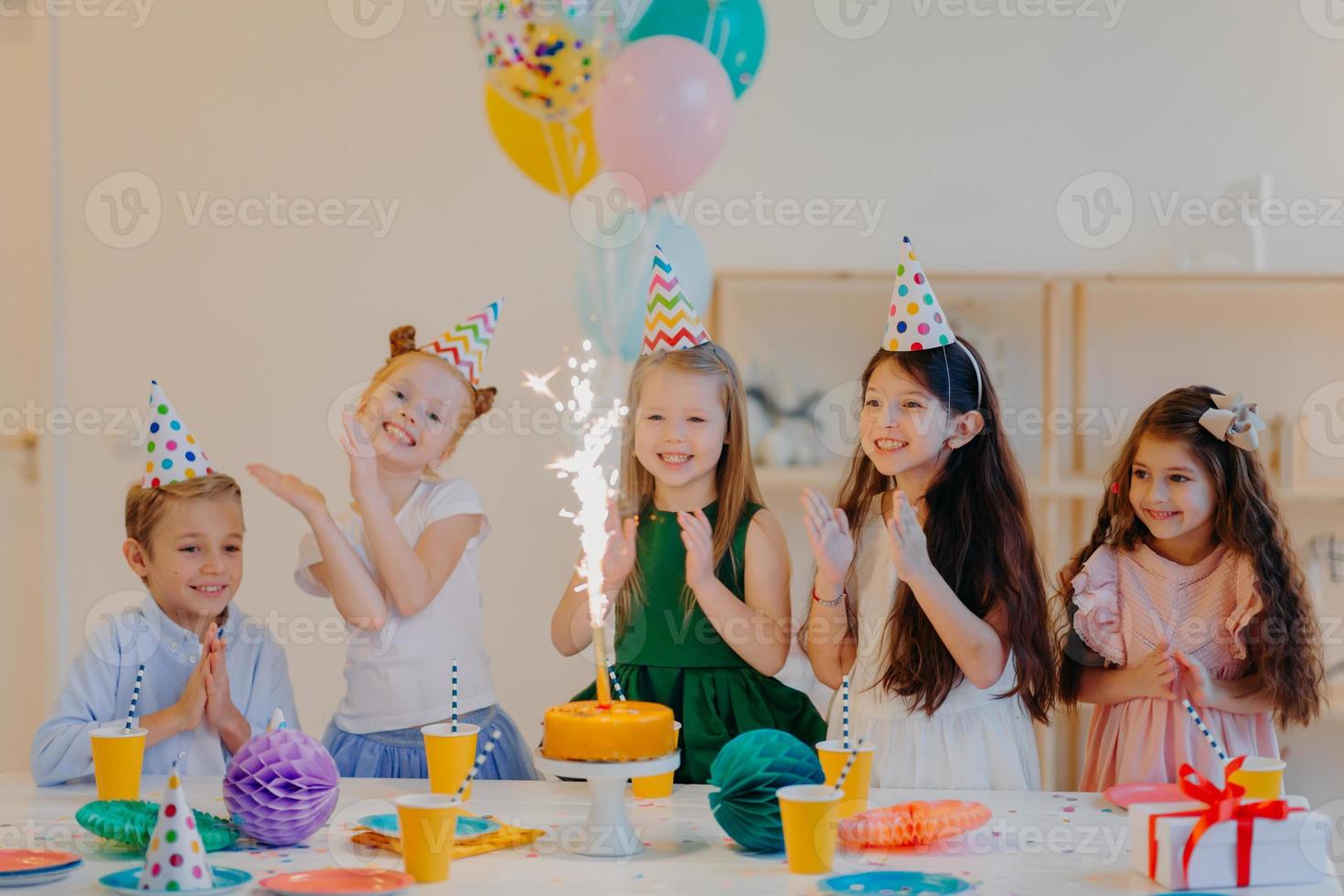 bambini e concetto di vacanza. felice che cinque amici guardino con gioia sulla torta con scintillio, festeggiano il compleanno, indossano cappelli a cono da festa e tengono palloncini ad aria, hanno espressioni felici foto