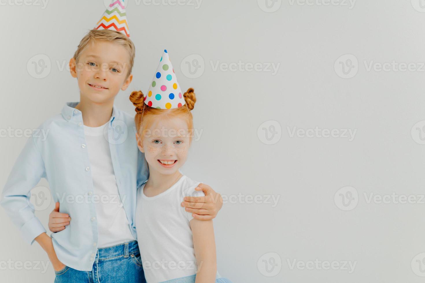 colpo isolato di fratello e sorella felici si coccolano, hanno espressioni positive, indossano cappelli da festa, vanno a festeggiare il compleanno, stanno su sfondo bianco, copiano lo spazio da parte. bambini allegri foto