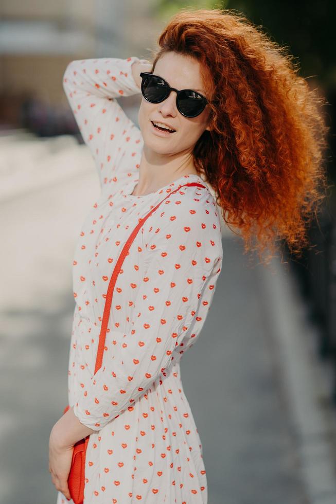 inquadratura verticale di bella donna con occhiali da sole alla moda, ha i capelli ricci e folti rossi, porta una piccola borsa, posa all'aperto su sfondo sfocato, fa una passeggiata in città, si gode le vacanze estive, esplora i luoghi d'interesse foto