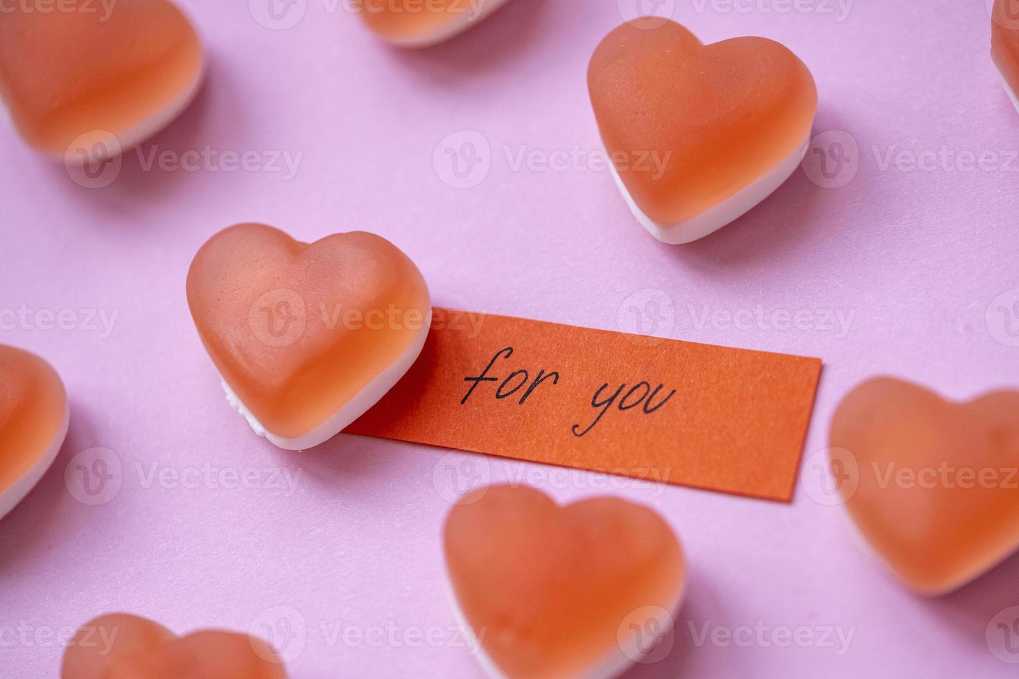marmellata di caramelle rosse a forma di cuore con una nota per te su sfondo rosa. caramelle gommose di zucchero. concetto di san valentino. motivo geometrico. design minimale, piatto, vista dall'alto. foto
